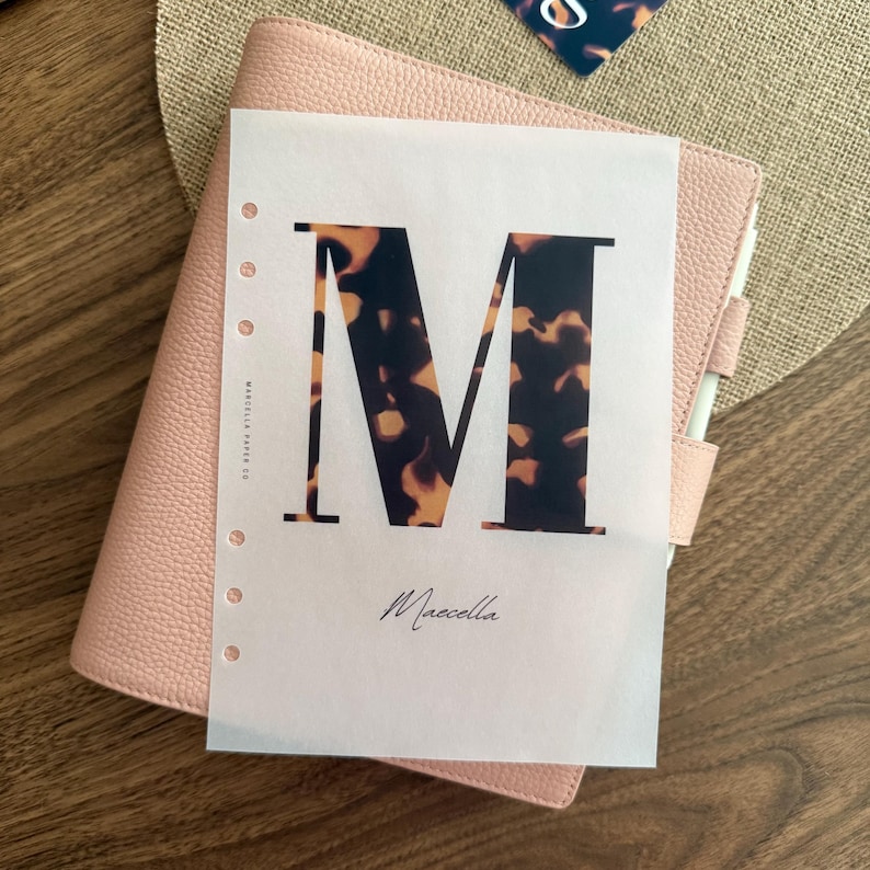 May include: A light pink textured planner with a white translucent insert page featuring a large letter M with a brown and black tortoiseshell pattern. The page includes the text Marcella Paper Co and the cursive name Maecella.