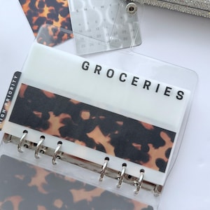 May include: Clear plastic binder with a tortoiseshell pattern and the word "GROCERIES" printed in black. A "HIGH PRIORITY" tab is visible. The binder is open, revealing the metal rings and additional inserts.