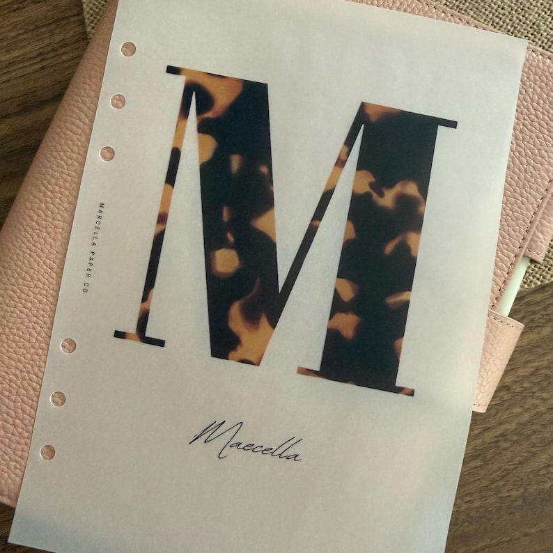 May include: A translucent planner insert page featuring a large letter M with a brown and black tortoiseshell pattern. The text Marcella Paper Co is printed vertically on the left side, and the name Maecella is written in script at the bottom center.