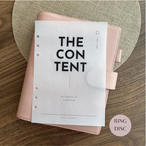 May include: A light pink textured leather ring binder planner with a translucent vellum cover sheet. The cover features black text reading THE CONTENT, 2040/2060, MY COMPLETE LIFE IN ONE SPACE, and MARCELLA PAPER CO, with six punched holes on the left.