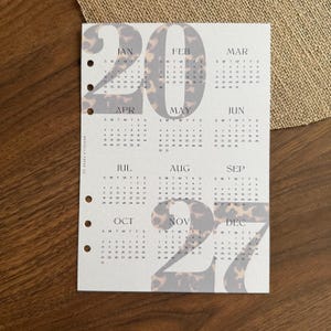 Könnte beinhalten: Ein weißes Kalenderblatt für das Jahr 2027 mit sechs Ringbuchlöchern auf der linken Seite. Die großen Ziffern 2027 sind mit einem braun-schwarzen Leopardenmuster überlagert. Der Text MARDELLA PAPER CO ist vertikal auf der linken Seite gedruckt.