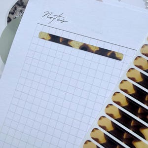 May include: A white notepad with a grid pattern and the word "Notes" in cursive. A decorative, rectangular sticker with a tortoiseshell pattern is adhered to the page. Several matching stickers are visible on a white backing.