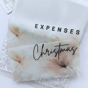 May include: Clear plastic organiser with two inserts. One insert reads "EXPENSES" in black block letters. The other insert reads "Christmas" in cursive script. Both inserts feature a floral design in neutral tones. A calendar is visible in the background.