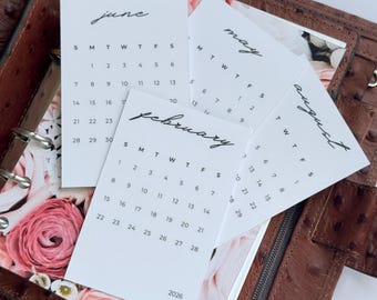 May include: A brown leather planner with small white calendars for June, May, February, and August. The calendars show the days of the week and the year 2026. The planner is open, revealing a floral design. The planner is made of leather.