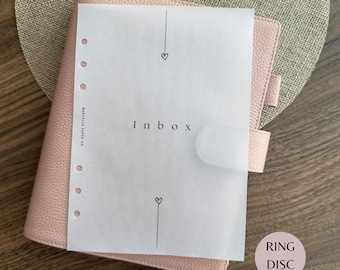 Puede incluir: Una agenda de cuero texturizado rosa claro con una hoja de papel vegetal translúcido encima. El papel muestra la palabra Inbox en el centro, flanqueada por dos líneas verticales y pequeños corazones. Seis perforaciones circulares visibles a la izquierda.