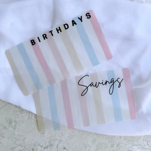 May include: Two clear, rectangular budget envelopes with pastel stripes. One reads "BIRTHDAYS" in black block letters, the other reads "Savings" in a cursive font. The envelopes are on a white surface.