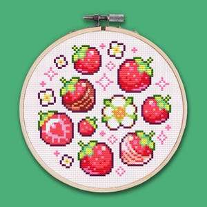 Puede incluir: Un bastidor de bordado de punto de cruz con un diseño de fresas rojas, flores blancas y estrellas rosas. Las fresas tienen copas verdes y están dispuestas en un patrón circular sobre un fondo de tela blanca. El bastidor de madera es visible.