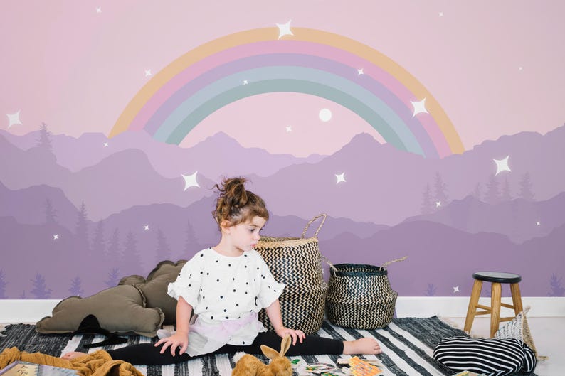 Pu&ograve; includere: Una cameretta con un murale raffigurante un arcobaleno, montagne e stelle nei toni del viola e del rosa. La stanza comprende cesti intrecciati, uno sgabello in legno e un tappeto a righe bianche e nere.