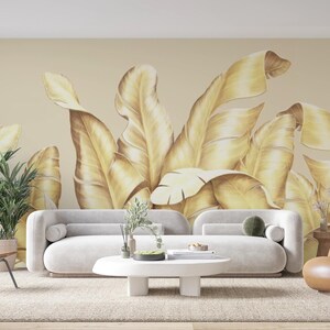 May include: A living room interior design featuring a white sofa and a brown leather armchair. The wall is decorated with a large-scale wallpaper featuring a pattern of golden banana leaves.