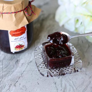 Op de afbeelding: Een glazen pot met Perzische kersenjam met een bruin papieren etiket en een rood touwtje om de deksel. De pot staat op een grijs oppervlak. Een klein glazen schaaltje met een doorzichtig deksel bevat meer kersenjam. Een lepel schept de jam uit het schaaltje.