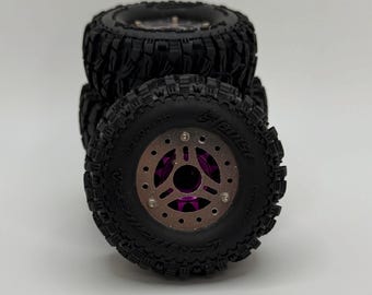 Pode incluir: Quatro pneus off-road pretos empilhados. Os pneus têm um padrão de piso profundo e são montados em aros roxos e prateados. Os pneus são projetados para veículos de controle remoto. O diâmetro é de aproximadamente 10 cm.