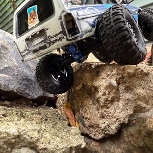 Pode incluir: Um ve&iacute;culo off-road de brinquedo branco e azul com pneus pretos grandes &eacute; mostrado escalando rochas. O ve&iacute;culo tem um acabamento de pintura desgastado e adesivos nas janelas. Os pneus s&atilde;o projetados para terrenos off-road.
