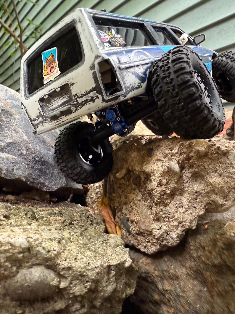 Pode incluir: Um ve&iacute;culo off-road de brinquedo branco e azul com pneus pretos grandes &eacute; mostrado escalando rochas. O ve&iacute;culo tem um acabamento de pintura desgastado e adesivos nas janelas. Os pneus s&atilde;o projetados para terrenos off-road.
