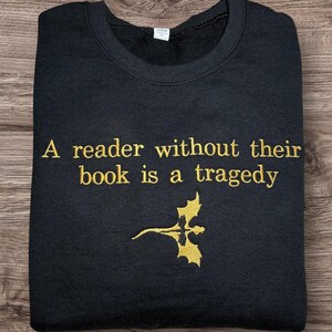 Op de afbeelding: Zwart sweatshirt met gouden tekst "A reader without their book is a tragedy" en een gouden draakafbeelding. Het sweatshirt is op een houten oppervlak gevouwen.