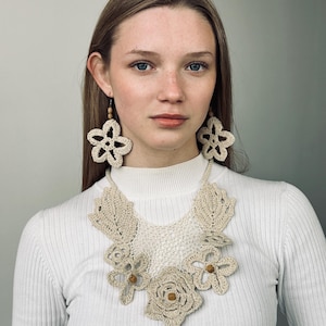 May include: A cream-colored crochet necklace and matching earrings. The necklace features floral and leaf designs with wooden bead accents. The earrings are large, openwork flower shapes. The jewelry is displayed on a person wearing a white ribbed long-sleeve shirt.