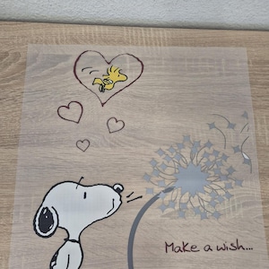 Peut inclure: Illustration de dessin animé avec Snoopy soufflant sur un pissenlit, avec un cœur contenant Woodstock au-dessus. La phrase « Make a wish... » est écrite sous le pissenlit. L'image est en noir, blanc, gris, jaune et rouge.