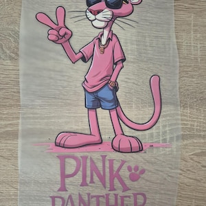 Peut inclure: Illustration du personnage de dessin animé Pink Panther portant des lunettes de soleil, une chemise rose, un short bleu et une chaîne en or, faisant le signe de la paix. Le texte "PINK PANTHER" est imprimé en rose en dessous.