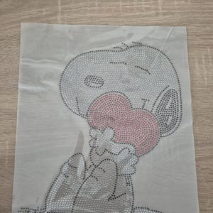 Puede incluir: Una lámina rectangular transparente con un diseño de pedrería de Snoopy sosteniendo un corazón rosa. El diseño es principalmente plateado con detalles negros, creando un efecto brillante. La imagen está sobre una superficie clara.