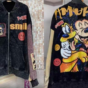 Puede incluir: Chaqueta vaquera negra con cremallera, con gráficos de Mickey Mouse y Goofy. La chaqueta tiene parches con las palabras "T+Tang" y "smile", y diseños de caritas sonrientes. Mangas beiges y moradas.