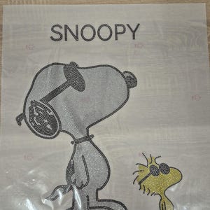 Puede incluir: Un diseño de Snoopy y Woodstock, creado con brillantes pedrería. La palabra "SNOOPY" está escrita con pedrería encima de la imagen. Snoopy lleva gafas de sol y Woodstock es amarillo con gafas de sol.