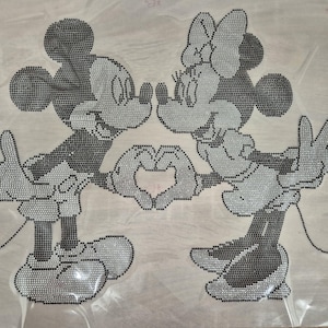 May include: A rhinestone design featuring Mickey Mouse and Minnie Mouse in a loving embrace. The figures are in black and grey, with Mickey and Minnie's hands forming a heart shape. The design is on a clear, textured backing.