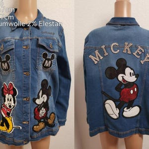 Może przedstawiać: Niebieska dżinsowa kurtka z naszywkami Mickey Mouse i Minnie Mouse. Kurtka ma naszywkę "MICKEY" z kryształkami na plecach.