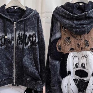 Könnte beinhalten: Ein dunkelgrauer Kapuzenpullover mit Reißverschluss und einem Used-Look-Effekt. Auf der Vorderseite steht der Text "RODEO MUSE" in Paillettenbuchstaben. Auf der Rückseite befindet sich ein Minnie Mouse-Design mit einer braunen Schleife und einem weißen Gesicht.
