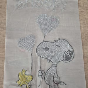 Puede incluir: Una tela gris claro con un diseño de pedrería de Snoopy y Woodstock. Snoopy sostiene dos globos en forma de corazón. La palabra "Snoopy" está escrita con pedrería en la parte superior de la tela.