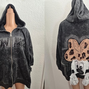 Könnte beinhalten: Ein anthrazitfarbener Kapuzenpullover mit Reißverschluss und Used-Waschung. Die Vorderseite zeigt einen paillettenbesetzten Text, und die Rückseite zeigt eine große Applikation einer Zeichentrickfigur mit Schleife und Handschuhen. Der Hoodie hat eine Kapuze mit Kordelzug und einen durchgehenden Reißverschluss.