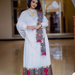 May include: A white dress with a colorful embroidered trim and a matching sash. The dress has a long, flowing skirt and a high neckline.