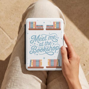 Può includere: Custodia bianca per e-reader con un design a pile di libri su scaffali e la scritta "Meet me at the Bookshop" in blu. La custodia è tenuta da una persona che indossa pantaloni beige.