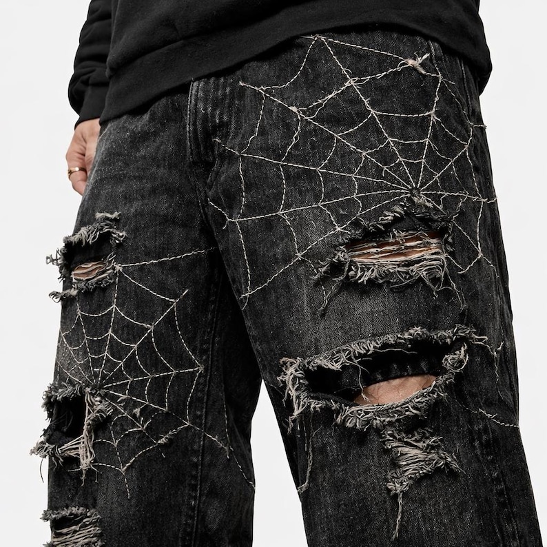 Pode incluir: Jeans de ganga pretos com detalhes desgastados e bordados de teia de aranha brancos. Os jeans apresentam m&uacute;ltiplas rasgaduras, criando uma est&eacute;tica grunge. O design da teia de aranha &eacute; costurado na frente.