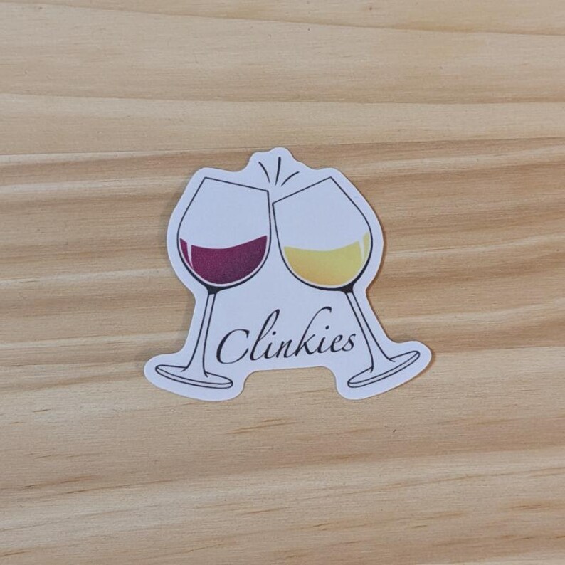 Puede incluir: Una pegatina blanca con dos copas de vino brindando. La copa de la izquierda est&aacute; llena de vino tinto y la de la derecha de vino blanco. El texto "Clinkies" est&aacute; escrito debajo de las copas.