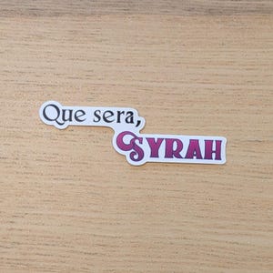 May include: A sticker with the text "Que sera, Syrah" in white lettering on a pink background.