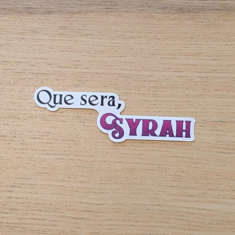 May include: A sticker with the text "Que sera, Syrah" in white lettering on a pink background.