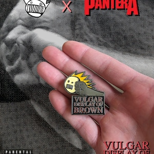 May include: A black and gold metal pin featuring a cartoon fist holding a microphone with the text "Vulgar Display of Brown" in red and black letters.