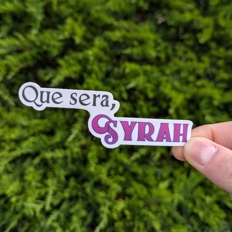 May include: A white sticker with the text "Que sera, SYRAH" in pink and purple letters. The sticker is held in a hand against a green background.