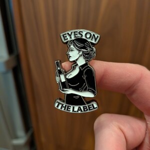 May include: Black and white enamel pin featuring a woman holding a wine bottle. The pin has the text "EYES ON THE LABEL" in a banner above and below the woman. The woman is wearing a black dress and has her hair up.