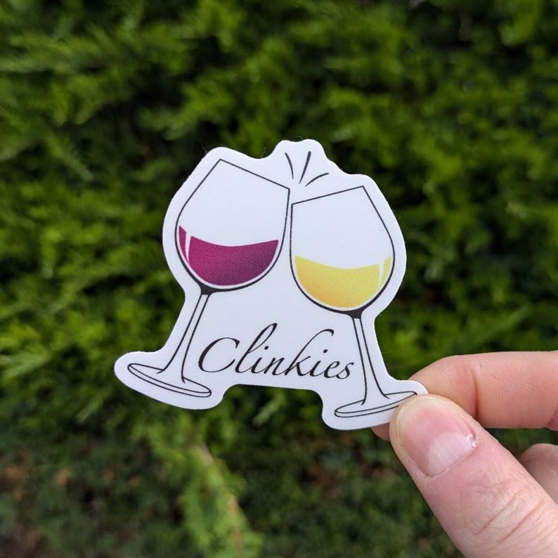 Puede incluir: Pegatina blanca con dos copas de vino brindando, una llena de vino tinto y la otra de vino blanco. El texto "Clinkies" est&aacute; escrito debajo de las copas.