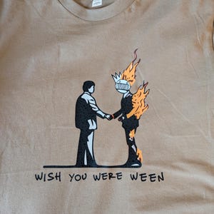 Puede incluir: Camiseta color beige con un diseño gráfico en blanco y negro de dos figuras dándose la mano, una de ellas en llamas. El texto "WISH YOU WERE WEEN" está impreso debajo. La camiseta está hecha de un material suave.