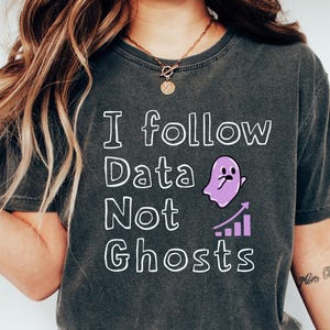 Puede incluir: Camiseta gris oscuro con el texto "I follow Data Not Ghosts" en blanco. También presenta un fantasma morado y un gráfico de barras. La camiseta es de estilo casual.