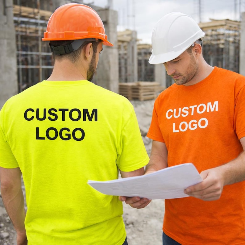 K&ouml;nnte beinhalten: Zwei Bauarbeiter tragen Helme und T-Shirts mit wei&szlig;em "CUSTOM LOGO" Aufdruck. Eines ist leuchtend gelb, das andere orange. Sie pr&uuml;fen Baupl&auml;ne auf einer Baustelle.