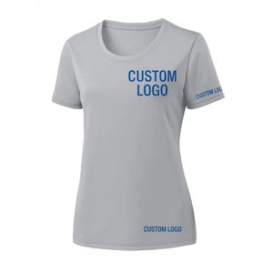 May include: A light gray short-sleeve athletic shirt with a crew neck. The words "CUSTOM LOGO" are printed in blue on the chest, sleeve, and lower front of the shirt. The shirt is made of a lightweight, breathable fabric.