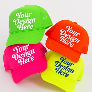 May include: Four brightly colored trucker hats stacked together. The hats are neon green, orange, hot pink, and yellow. Each hat has the text "Your Design Here" in white or blue script. The hats have a mesh back and a curved brim.
