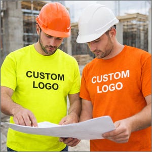 K&ouml;nnte beinhalten: Zwei Bauarbeiter tragen Helme und T-Shirts mit dem Text "CUSTOM LOGO". Ein Arbeiter tr&auml;gt einen orangefarbenen Helm und ein gelbes T-Shirt. Der andere Arbeiter tr&auml;gt einen wei&szlig;en Helm und ein orangefarbenes T-Shirt. Sie betrachten einen Bauplan.