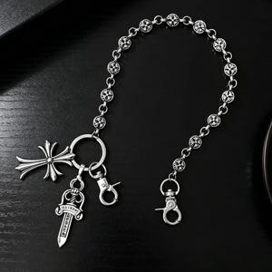 Könnte beinhalten: Silberfarbene Kette mit dekorativen Anhängern. Die Kette hat kreisförmige Glieder mit kreuzförmigen Akzenten. Zu den Anhängern gehören ein Kreuz, ein Dolch und zwei Karabinerverschlüsse. Die Kette ist für die Befestigung an einer Gürtelschlaufe oder Tasche konzipiert.