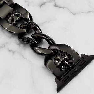 以下が含まれることがあります： 黒い時計バンド、チェーンリンクのデザイン。リンクには花のモチーフが飾られています。バンドは光沢のある暗い素材でできており、白い大理石の背景に配置されています。