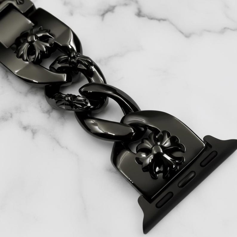 以下が含まれることがあります： 黒い時計バンド、チェーンリンクのデザイン。リンクには花のモチーフが飾られています。バンドは光沢のある暗い素材でできており、白い大理石の背景に配置されています。