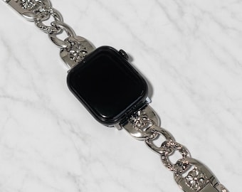 May include: A silver-toned watch band with a chain-link design. The band is attached to a black smartwatch. The links have a decorative floral pattern. The watch band is made of metal.