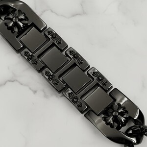以下が含まれることがあります： 黒いブレスレットのクローズアップ。フローラルデザイン。ブレスレットは長方形と正方形のリンクでできており、各リンクに装飾的なフローラル要素があります。ブレスレットは白とグレーの大理石の表面にあります。